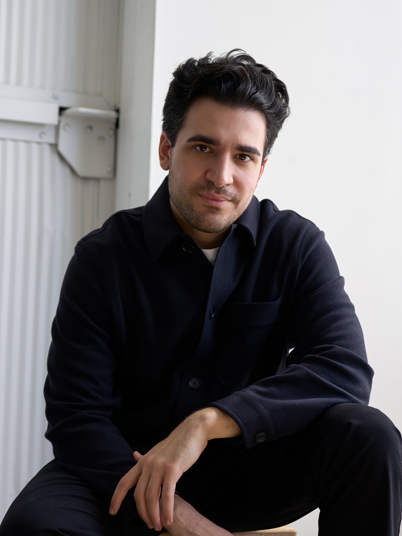 Samvel Krajian in a dark overshirt, seated with one arm resting across his knee, looking at the camera.
